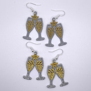 May include: A pair of silver earrings with gold champagne glass charms. The glasses are filled with gold glitter and have a gold star on top.