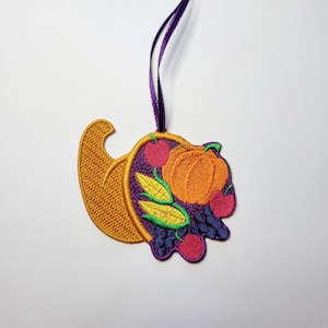 Puede incluir: Adorno bordado de cornucopia con una cinta morada. La decoración con temática de cosecha presenta un cuerno dorado lleno de calabaza, manzanas, uvas y maíz. El adorno está hecho con hilo naranja, morado, verde y rojo.