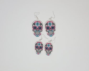 May include: A pair of silver earrings with white sugar skull designs. The skulls have pink and blue floral details and black eyes.