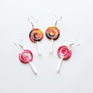 Pode incluir: Três pares de brincos de pirulito coloridos. Os brincos são feitos de fio e têm um palito branco. Um par é vermelho e rosa, um par é laranja, amarelo e preto, e o outro par é vermelho e rosa.
