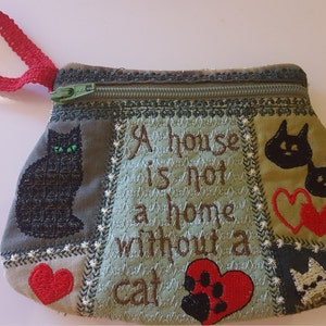 May include: A green fabric zippered pouch with a red handle. The pouch is decorated with embroidered black cats, red hearts, and the text "A house is not a home without a cat".