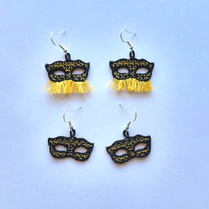 May include: Four pairs of black masquerade mask earrings with yellow fringe. The masks have a geometric pattern.