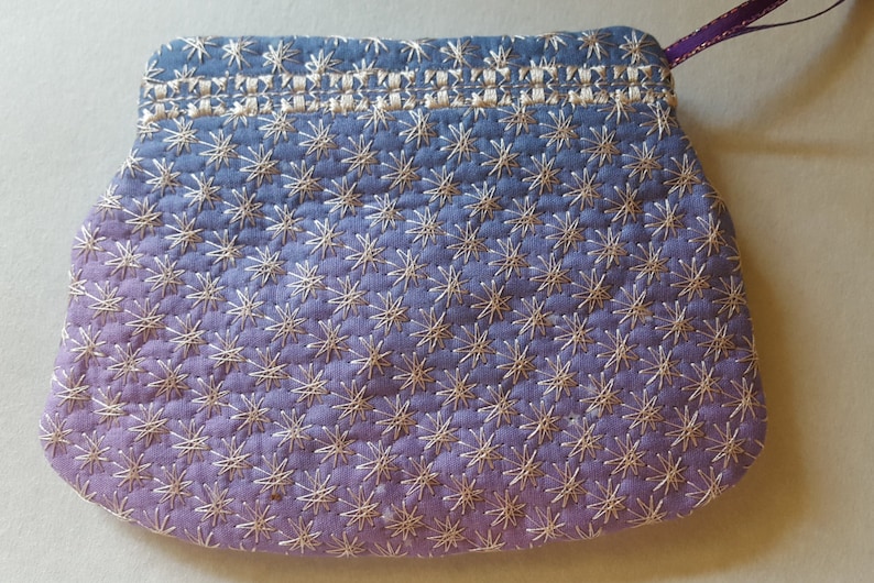 May include: A small, purple and blue fabric pouch with a white star pattern. The pouch has a drawstring closure and a purple ribbon.