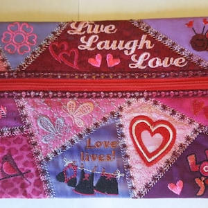 Può includere: Una pochette con cerniera rosa e rossa con un design a patchwork che presenta cuori, uccelli, fiori e il testo "Live Laugh Love" ricamato.