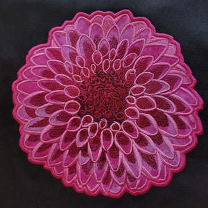 Pode incluir: Um remendo bordado com forma de flor circular, de cor rosa e vermelha. A flor tem um design em camadas com várias pétalas.