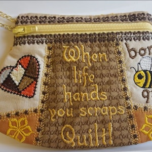 May include: A beige fabric pouch with a zipper closure and a gold chain. The pouch is decorated with embroidered designs, including a heart, a bee, and the text "When life hands you scraps Quilt!"