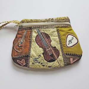 May include: A small, patchwork coin purse with musical instrument embroidery. The purse features a guitar, violin, and treble clef. It has a gold zipper and a matching wrist strap. The color palette includes brown, green, and gold.