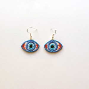 May include: A pair of silver earrings with blue, white and red embroidered evil eye charms.
