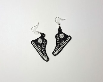 Pode incluir: Um par de brincos pretos bordados em forma de tênis de cano alto com costuras brancas e um círculo branco na lateral.