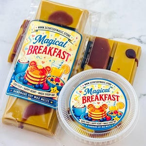 May include: A rectangular bar and a round container of wax melts, both labeled Magical Breakfast. The labels feature illustrations of pancakes, a drink, and cereal. The wax is a golden yellow color with dark brown accents, packaged in clear plastic.