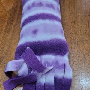 May include: A purple and white fleece cat toy. The cylindrical toy has a tie-dye pattern with shades of purple and white. The bottom of the toy is fringed with strips of purple fleece. The toy is designed for cats.
