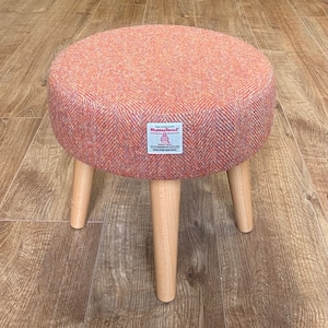 May include: A round orange and white tweed footstool with wooden legs. The footstool has a label that reads "Habia Tweed".