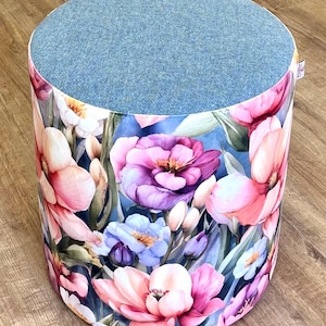May include: A round ottoman with a blue felt top and a floral print base. The base features large pink, purple, and white flowers against a blue background. The ottoman is a decorative furniture piece.