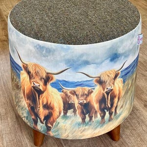 May include: Round ottoman with a brown tweed top and a printed fabric base featuring a herd of Highland cattle in a grassy field under a blue sky. The ottoman has three wooden legs.