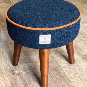 以下が含まれることがあります： オレンジ色のトリムが付いた、丸いネイビーブルーの布張りのスツール。スツールには3本の木製の脚が付いています。生地はヘリンボーン柄です。スツールの直径は約30cmです。