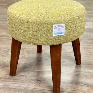 Può includere: Uno sgabello rotondo in tweed giallo con tre gambe in legno. Lo sgabello ha un'etichetta che recita "Harris Tweed".