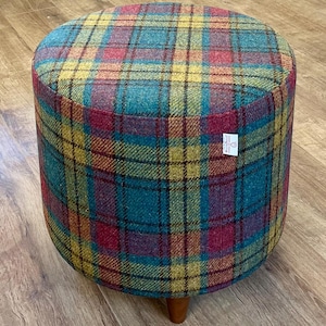 May include: A round upholstered footstool with a colorful plaid pattern in teal, yellow, red, and purple. The stool has a cylindrical shape and rests on three wooden legs. A small tag is visible on the side.