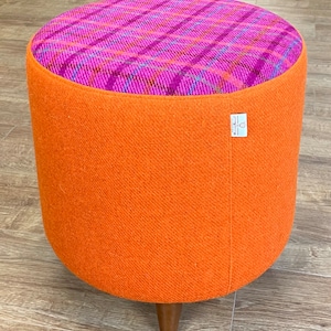 May include: A round orange and pink patterned footstool with wooden legs. The top of the footstool is covered in a pink and purple plaid fabric.