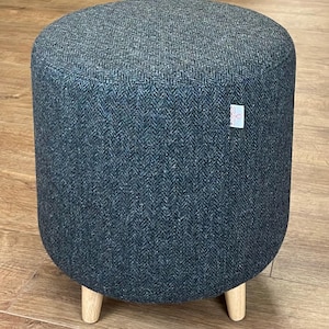 May include: A round, dark gray upholstered ottoman with a herringbone pattern. The ottoman has three short, light-colored wooden legs. The fabric has a textured appearance. A small tag is attached to the side.