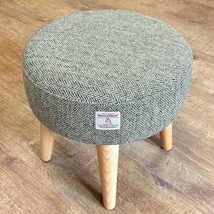 May include: A round footstool featuring a green and white herringbone patterned fabric upholstery. The stool has three light-coloured wooden legs and a white rectangular label on the side that reads Harris Tweed Hand Woven Cloth 100% Pure New Wool.