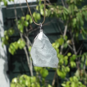 Puede incluir: Colgante de cristal azul claro de forma irregular, con una tapa y un asa de metal plateado, suspendido de un fino collar de cordón marrón. El fondo presenta follaje verde y un vistazo a un edificio.