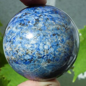May include: A polished, spherical lapis lazuli gemstone. The stone is a deep blue color with white and gold flecks, and a smooth, reflective surface. The gemstone is held between two fingers.