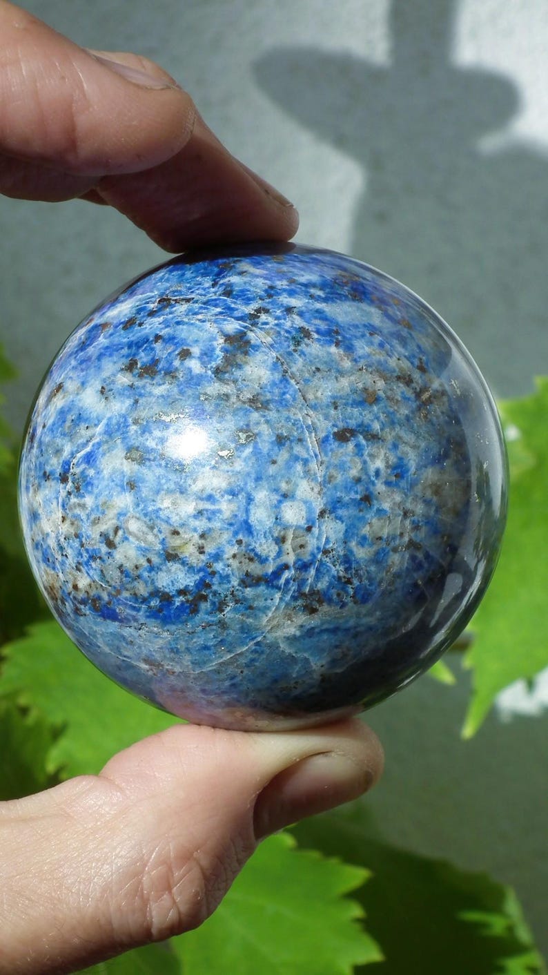 May include: A polished, spherical lapis lazuli gemstone. The stone is a deep blue color with white and gold flecks, and a smooth, reflective surface. The gemstone is held between two fingers.