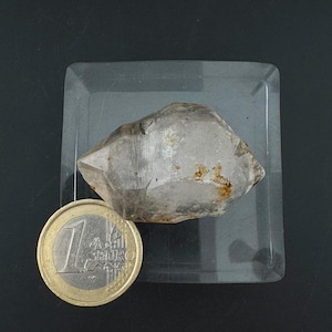 Pode incluir: Um cristal de quartzo bruto e translúcido com facetas naturais visíveis e pequenas inclusões minerais castanhas. O cristal está posicionado ao lado de uma moeda de 1 euro para escala, com um diâmetro de 23,25 mm, sobre uma base transparente.
