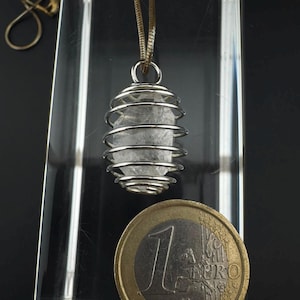 Może przedstawiać: Przezroczysty wisiorek z surowego kryształu zamknięty w srebrzystej metalowej spiralnej klatce, zawieszony na cienkim łańcuszku. Obok wisiorka umieszczono monetę jednoeurową, aby zapewnić wizualne odniesienie do rozmiaru i skali kryształowej klatki.