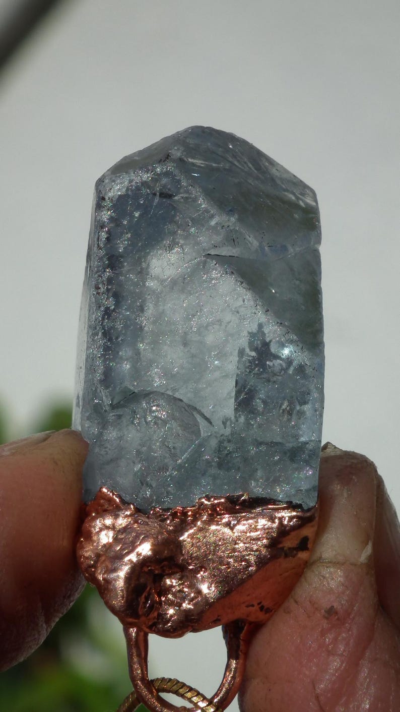 K&ouml;nnte beinhalten: Ein durchscheinender, hellblauer Kristallpunkt mit einer rauen, texturierten Kupferbasis. Der Kristall weist interne Br&uuml;che und Facetten auf, w&auml;hrend die metallische Kupferfassung an der Unterseite eine raue, unebene Oberfl&auml;che besitzt.