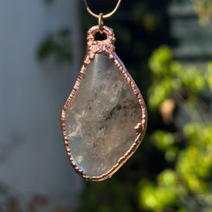 May include: A teardrop-shaped clear quartz pendant encased in a textured copper frame. The stone features natural internal inclusions and a translucent appearance. The pendant hangs from a thin metallic chain attached to a small copper loop at the top.