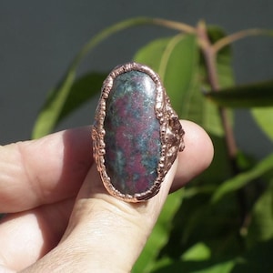 Può includere: Anello in rame con una pietra ovale dai toni rossi e verdi. La pietra è incastonata in una fascia di rame strutturata, creando un gioiello rustico e unico. L'anello è un accessorio di tendenza.
