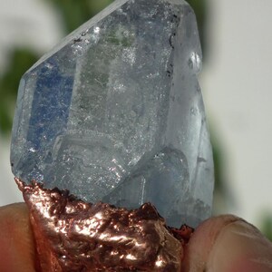 K&ouml;nnte beinhalten: Ein durchscheinender, hellblauer Kristallspitzen-Anh&auml;nger mit einer rauen, texturierten, galvanisierten Kupferbasis. Der Kristall hat eine facettierte, geometrische Form mit sichtbaren Einschl&uuml;ssen und eine metallische Kupferschlaufe unten.