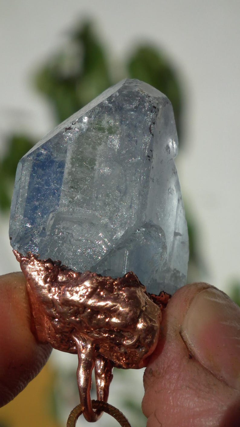 K&ouml;nnte beinhalten: Ein durchscheinender, hellblauer Kristallspitzen-Anh&auml;nger mit einer rauen, texturierten, galvanisierten Kupferbasis. Der Kristall hat eine facettierte, geometrische Form mit sichtbaren Einschl&uuml;ssen und eine metallische Kupferschlaufe unten.