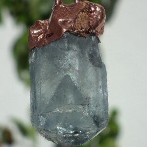 K&ouml;nnte beinhalten: Ein roher, durchscheinender hellblauer Kristallanh&auml;nger, eingefasst in eine strukturierte Kupferkappe. Der Kristall hat eine hexagonale, s&auml;ulenartige Form mit sichtbaren inneren Br&uuml;chen und lichtreflektierenden Facetten, h&auml;ngend an einer d&uuml;nnen Kette.