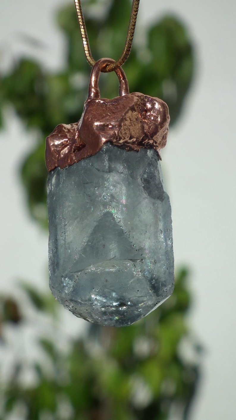 K&ouml;nnte beinhalten: Ein roher, durchscheinender hellblauer Kristallanh&auml;nger, eingefasst in eine strukturierte Kupferkappe. Der Kristall hat eine hexagonale, s&auml;ulenartige Form mit sichtbaren inneren Br&uuml;chen und lichtreflektierenden Facetten, h&auml;ngend an einer d&uuml;nnen Kette.