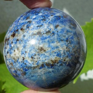May include: A blue sphere with a marbled pattern of white, brown, and darker blue. The polished stone is held between two fingers, showcasing its smooth, rounded form. The sphere's surface reflects light, highlighting its intricate veining.