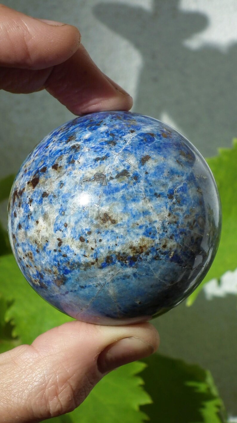 May include: A blue sphere with a marbled pattern of white, brown, and darker blue. The polished stone is held between two fingers, showcasing its smooth, rounded form. The sphere's surface reflects light, highlighting its intricate veining.