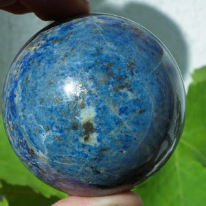 May include: A polished, spherical lapis lazuli gemstone. The deep blue stone has a marbled appearance with flecks of gold and white. The sphere is held between two fingers, showcasing its smooth, reflective surface. The background is blurred.