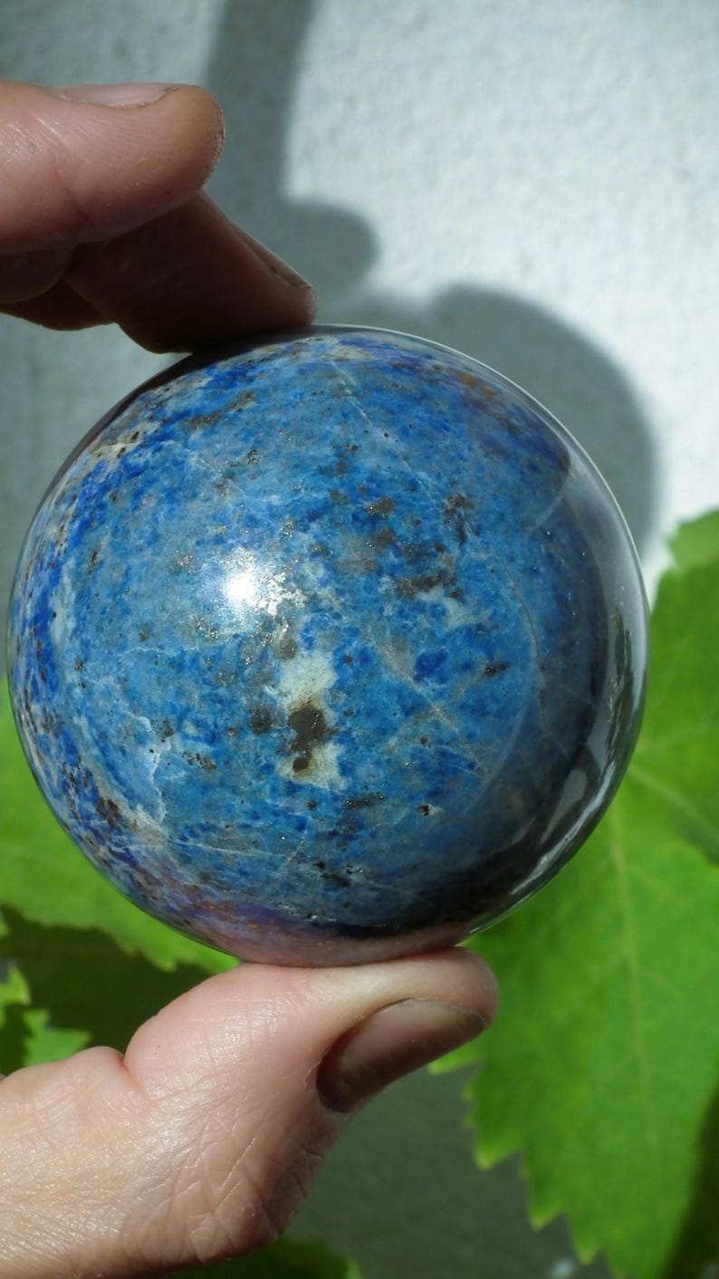 May include: A polished, spherical lapis lazuli gemstone. The deep blue stone has a marbled appearance with flecks of gold and white. The sphere is held between two fingers, showcasing its smooth, reflective surface. The background is blurred.