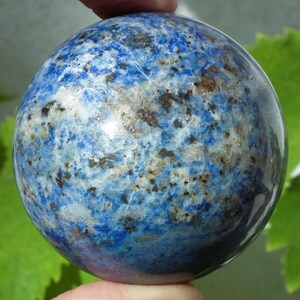 May include: A polished sphere of lapis lazuli, showcasing a deep blue hue with white and gold veining. The gemstone ball is held between fingers, with a blurred green background.