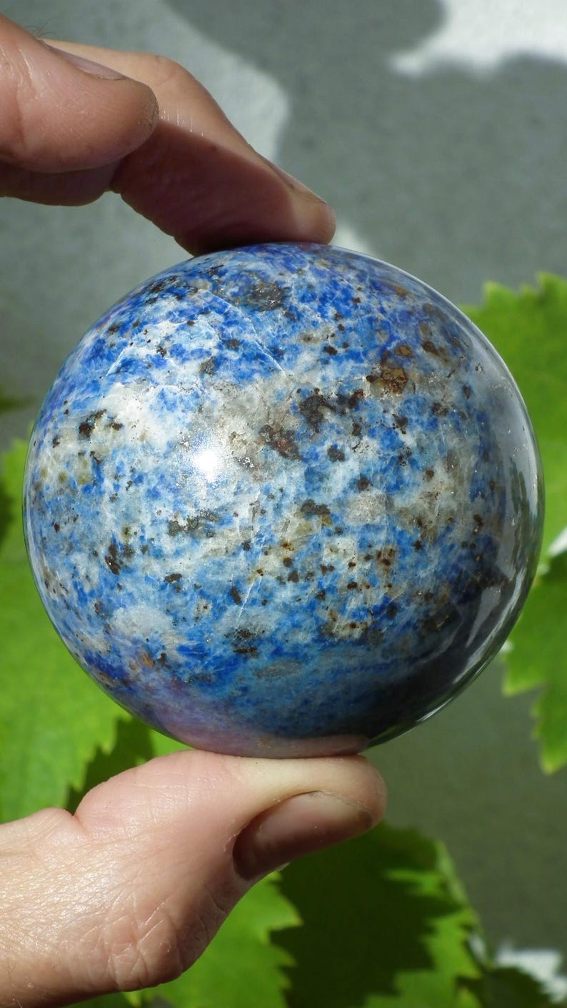 May include: A polished sphere of lapis lazuli, showcasing a deep blue hue with white and gold veining. The gemstone ball is held between fingers, with a blurred green background.
