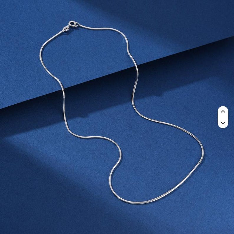 K&ouml;nnte beinhalten: Eine silberfarbene Schlangenkette mit einer glatten, flexiblen Metallkonstruktion und einem runden Federringverschluss am oberen Ende. Die Kette besteht aus eng miteinander verbundenen Gliedern, die ein durchgehendes, flie&szlig;endes Erscheinungsbild erzeugen.