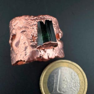 Könnte beinhalten: Ein breiter Kupferring mit einer texturierten, unebenen Oberfläche und einem einzelnen, vertikal eingesetzten dunkelgrünen Turmalinkristall. Der Ring liegt neben einer Ein-Euro-Münze mit einem Durchmesser von etwa 23,25 Millimetern.