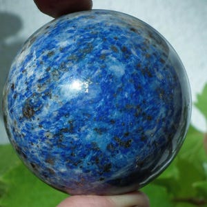 May include: A polished, spherical lapis lazuli gemstone. The deep blue stone features swirling patterns of white and flecks of pyrite, creating a marbled effect. The sphere is held in a hand.