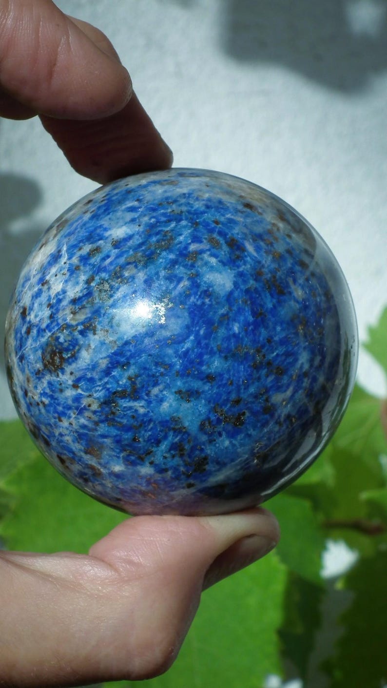 May include: A polished, spherical lapis lazuli gemstone. The deep blue stone features swirling patterns of white and flecks of pyrite, creating a marbled effect. The sphere is held in a hand.
