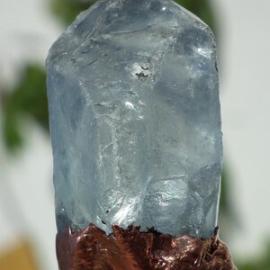 K&ouml;nnte beinhalten: Ein roher, durchscheinender hellblauer Kristallpunkt mit einer rauen, strukturierten kupferfarbenen Metallbasis. Der Kristall hat eine hexagonale Form mit nat&uuml;rlichen Facetten und Einschl&uuml;ssen, w&auml;hrend der metallische Boden dunkel und uneben ist.