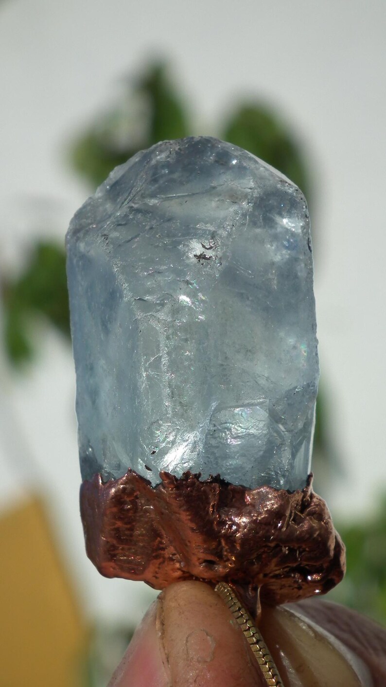 K&ouml;nnte beinhalten: Ein roher, durchscheinender hellblauer Kristallpunkt mit einer rauen, strukturierten kupferfarbenen Metallbasis. Der Kristall hat eine hexagonale Form mit nat&uuml;rlichen Facetten und Einschl&uuml;ssen, w&auml;hrend der metallische Boden dunkel und uneben ist.