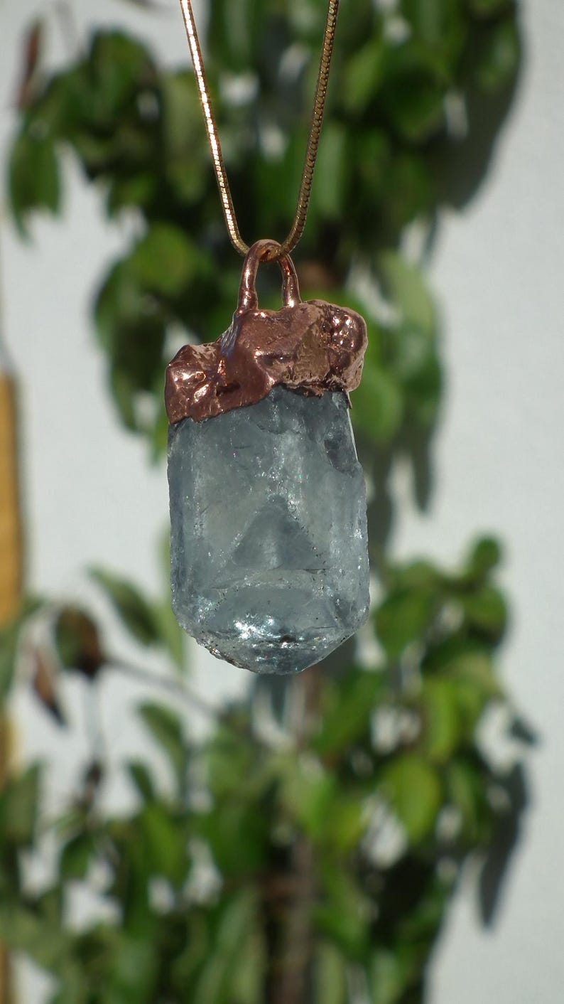 K&ouml;nnte beinhalten: Eine Anh&auml;ngerkette mit einem rohen, durchscheinenden hellblauen Kristallstein, der mit einer strukturierten, metallischen Kupferfassung versehen ist. Der Anh&auml;nger h&auml;ngt an einer d&uuml;nnen, passenden kupferfarbenen Kette und zeigt eine nat&uuml;rliche Optik.