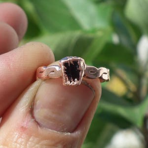 Op de afbeelding: Een koperkleurige ring met een gedraaide metalen band en een centrale zetting met een donkere, grillige zwarte steen. Het metaal heeft een warme metallic afwerking en de steen heeft een ruwe, onregelmatige textuur met een diepzwarte kleur.